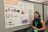 A biology student stands in front of a poster featuring information on bees and other pollinators