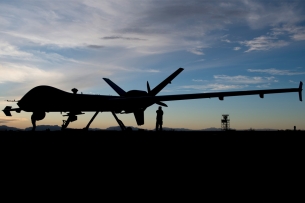 A silhouette of a military drone that looks like an airplane. The silhouette is set against a light blue sky with white clouds. A serviceperson standing beneath the rear of the drone, is also silhouetted.