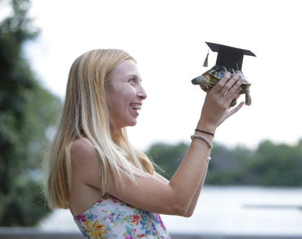 A student holds a turtle at eye level. The turtle is wearing a graduation cap on its shell.