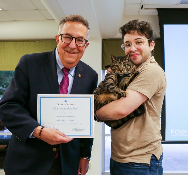 A student holds a cat while posing with an administrator who is awarding the cat a diploma from Eckerd College